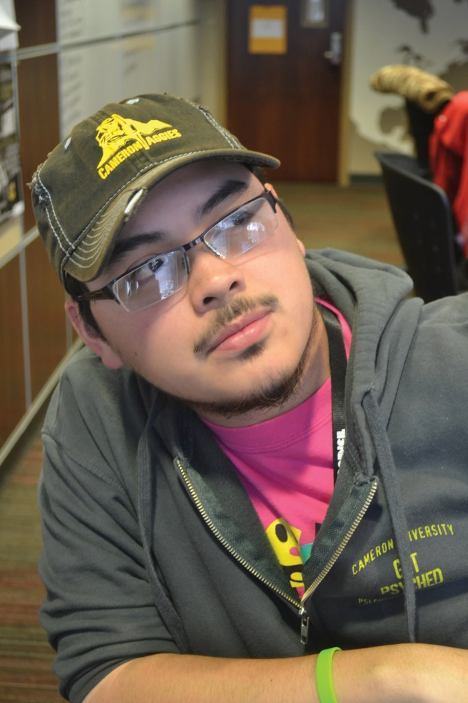 Older photo of Jacob Jardel with short hair in the Cameron Collegian newsroom wearing a Cameron Aggies baseball cap, a pink Cameron University Programming Activities Council t-shirt, and a grey Cameron University Psi Chi and Psychology Club zip hoodie