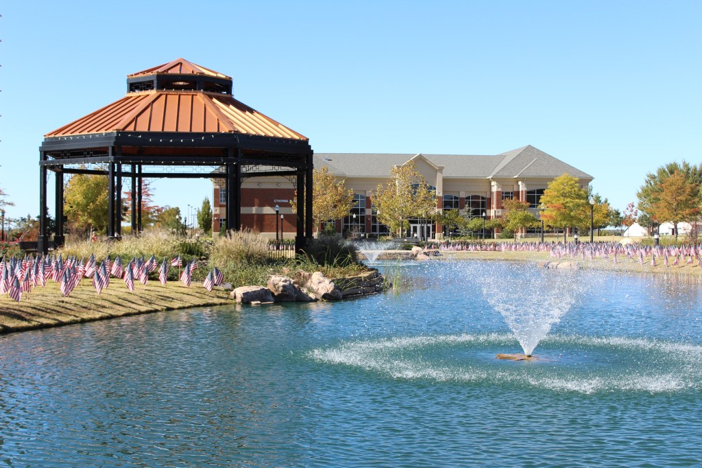 Photo from Cameron Lake with a visual of the gazebo on the left side, Cynthia Ross Hall in the background, and two fountains--one in the distance and one up close--with small American flags in the grass surrounding the lake and gazebo.