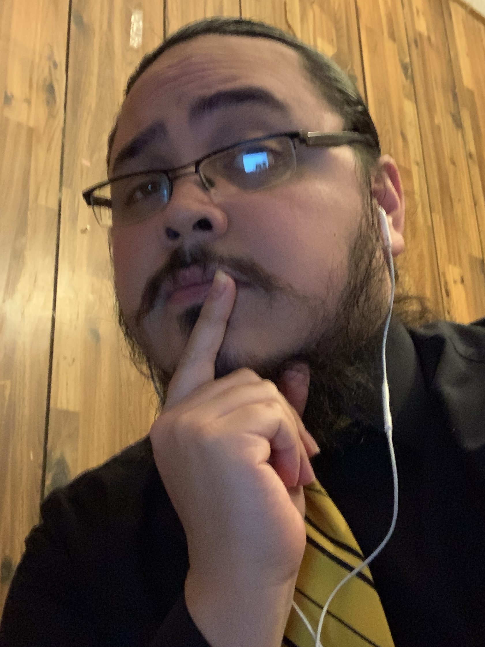 Photo of Jacob Jardel with glasses and his hair pulled back. He's wearing a black dress shirt with a yellow tie with black stripes, and he's wearing white Apple earbuds.