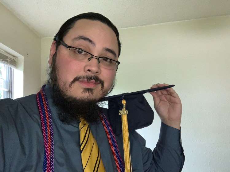 Photo of Jacob Jardel with glasses and hair tied back. He's wearing a blue-grey shirt and yellow tie with black stripes, and over his shoulders are graduation cords in magenta, blue, orange, and lighter blue. He is holding a black graduation cap with a yellow tassel that has the number 2020 on it.